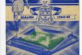 Sinivalkoisella pohjalla piirroskuva otteluohjelman tiedoin varustettuna, keskellä kuva jalkapallostadionista ja ylhäällä pelaaja jalkapallo kainalossaan.