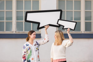 Kaksi naista katsoo toisiaan ja pitelee ylhäällä käsissään puhekuplia.