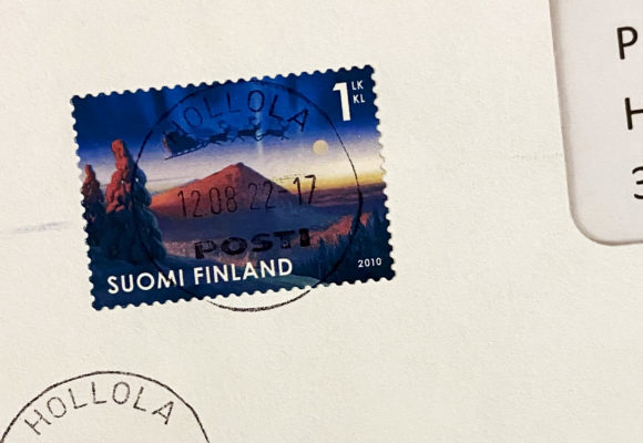 Kirjekuoressa leima päällään olevassa Suomen postimerkissä on etualalla lumen peittämiä kuusia vaarojen lähellä, taivaalla lentää poroja rekeä vetäen.
