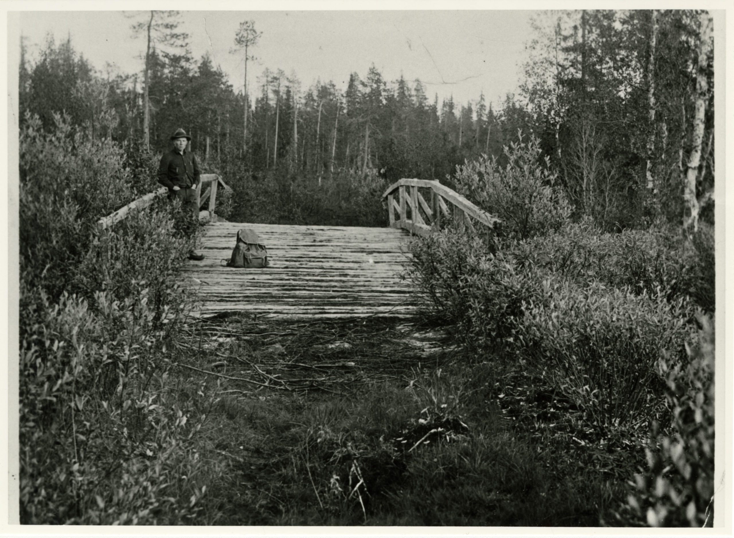 Mustavalkoisessa valokuvassa mies laukun kanssa puisella leveällä sillalla, ympärillä metsää.