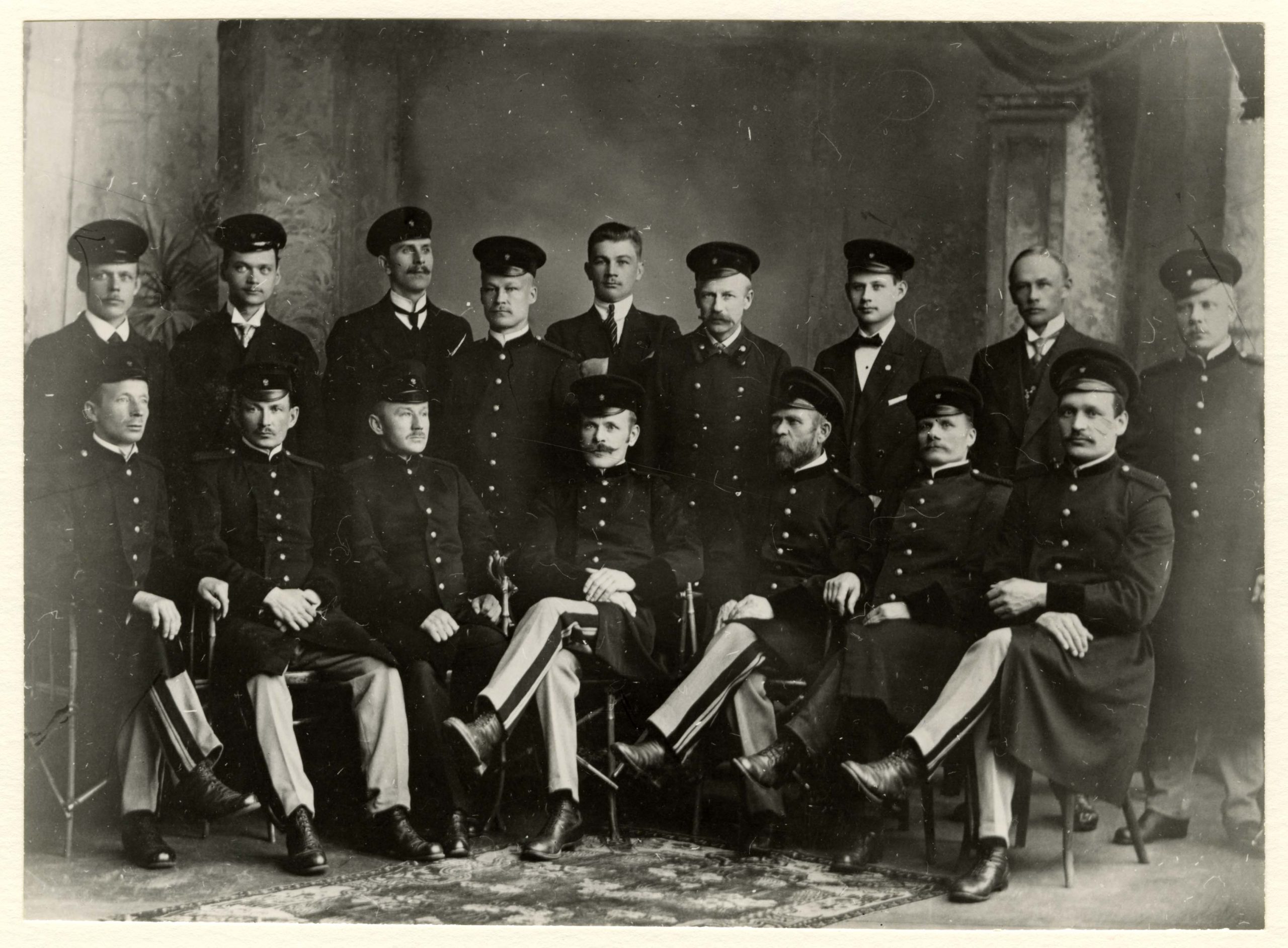Mustavalkoisessa kuvassa miehiä virkapuvuissa kahdessa rivissä. Ensimmäisessä rivissä olevat miehet istuvat tuoleilla, takarivissä seisovat.