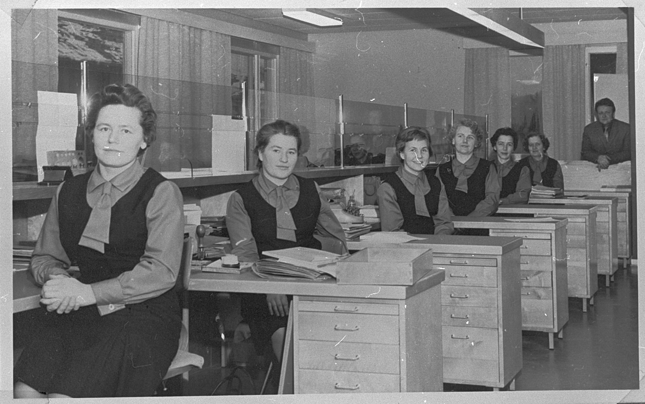 Mustavalkoisessa kuvassa samanlaisiin asuihin pukeutuneita naisia postitoimiston asiakaspalvelupisteiden luona. Taustalla pukuun pukeutunut mies.
