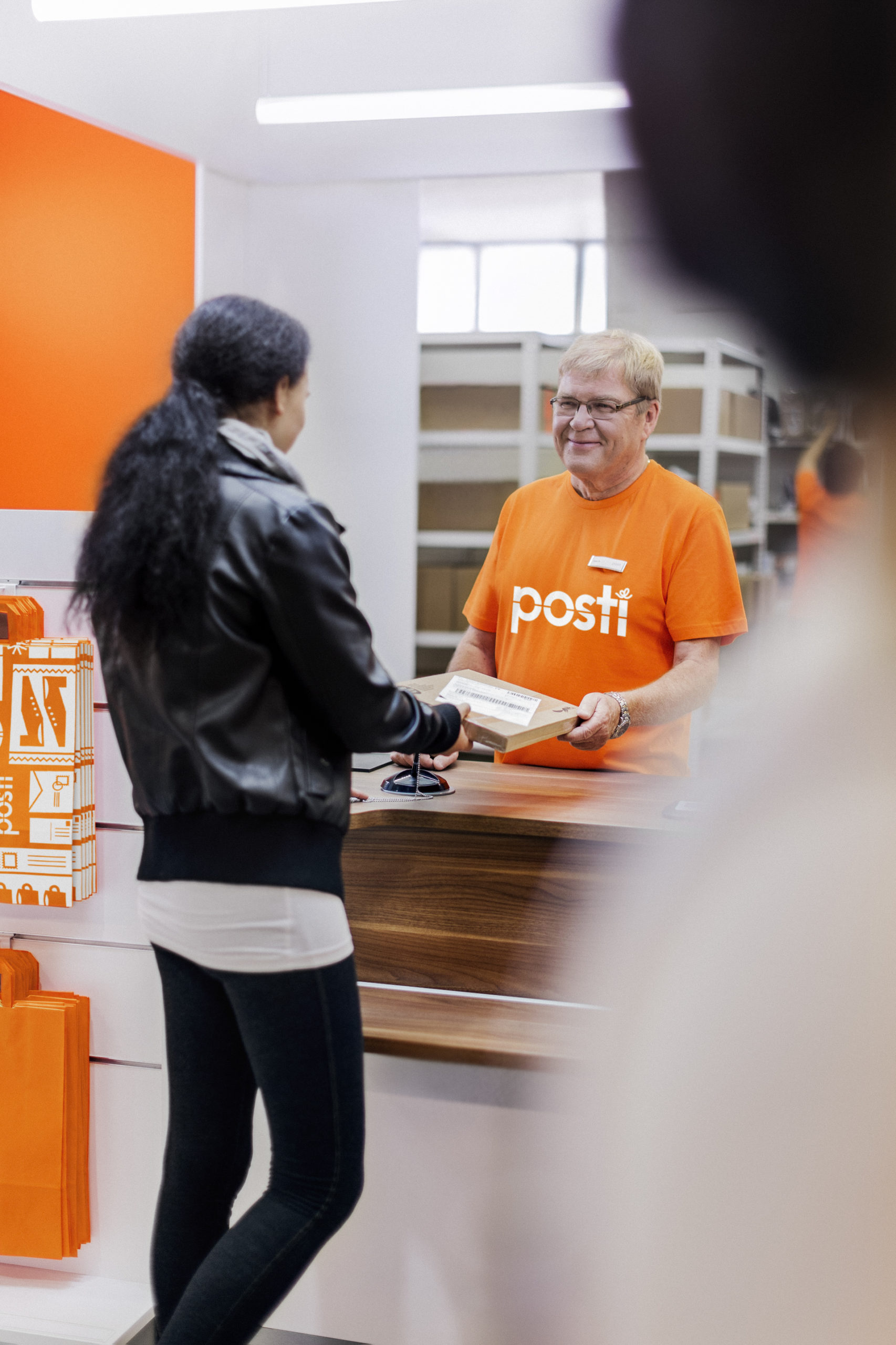 Asiakas hakemassa pientä pakettia Postista. Postityöntekijä antamassa lähetystä oranssissa t-paidassa, jossa Posti-logo.