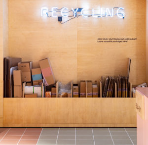 Recycling-valokyltin alapuolelta erilaisia käytettyjä postipaketteja koon mukaan jaoteltuina, osa taiteltuina puisessa ja lokeroidussa kulmauksessa.