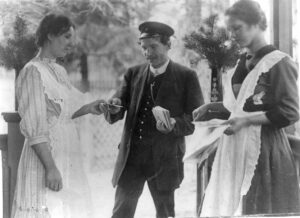 Posteljooni ojentaa kirjeitä kahdelle naiselle mustavalkoisessa kuvassa.