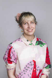 In a white patterned collared shirt, a blond-haired woman with a bun smiles in the staff photo.