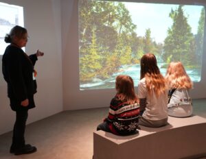 Opas esittelemässä seinälle heijastettua maisemapostikorttia penkillä istuville henkilöille.