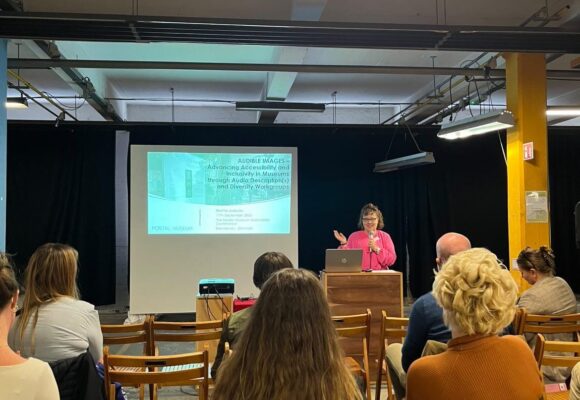 Etualalla ihmisiä istumassa kasvot eteenpäin. He katsovat vaaleanpunaiseen asuun pukeutunutta henkilöä, jolla on mikrofoni kädessä. Taustalla on diaesitys.