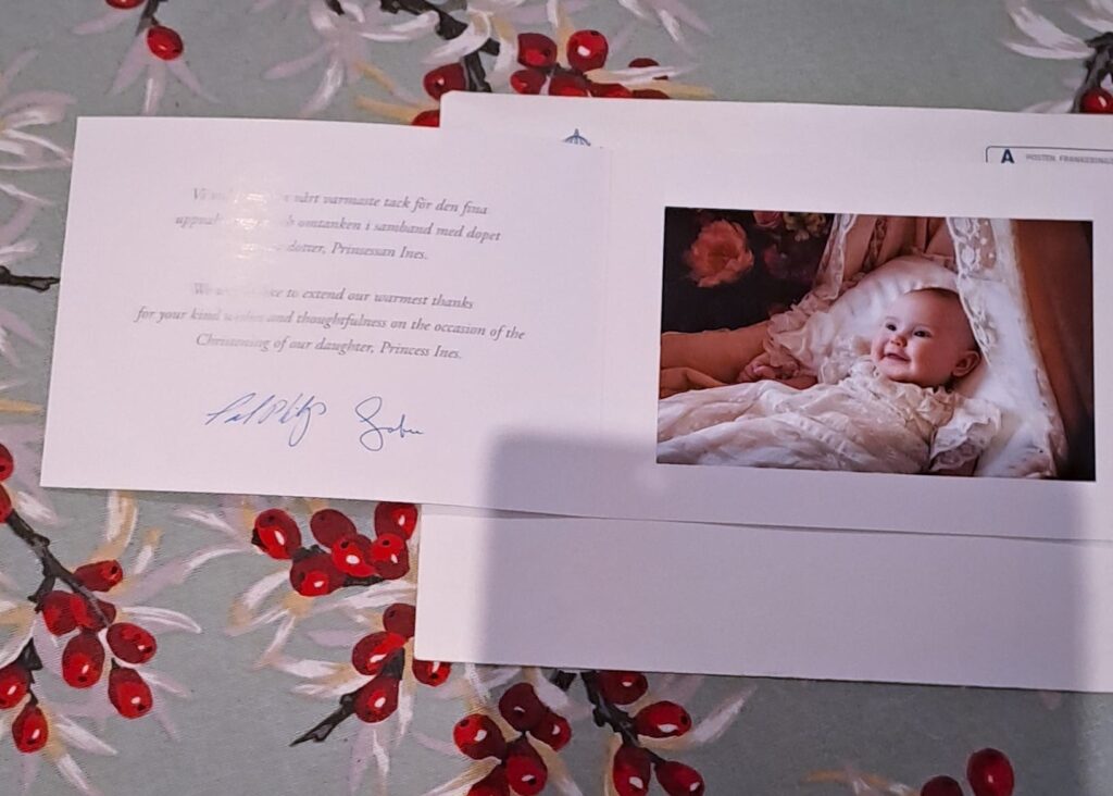 Vaakatasoinen taitettu kortti kirjekuoren ja kuviollisen pöytäliinan päällä pöydällä. kortin oikealla puolella on hymyilevä vauva kastemekossa kehdossa. Vasemmalla puolella on ruotsiksi ja englanniksi painettua tekstiä sekä kaksi allekirjoitusta.