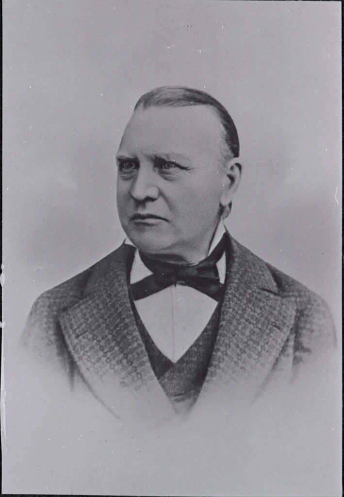 Mustavalkoisessa kuvassa on mieshenkilö puvun takissa ja rusetti kaulassa.