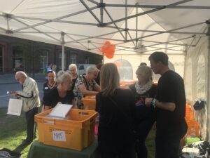 ihmisiä teltan alla ulkona, pöydillä oranssit laatikot.