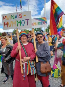 Kaksi ihmistä pukeutuneina vanhoihin vaatteisiin Pride-marssilla kyltit kädessään.