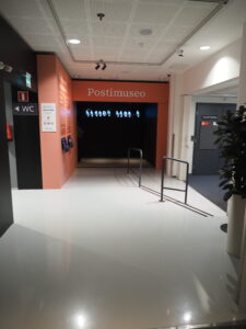 Käytävä, joka päättyy vaaleanpunaiseen sisäänkäyntiin. Ylhäällä teksti Postimuseo. Käytävän päässä mustaa ja kyyhkysen kuvia seinällä.