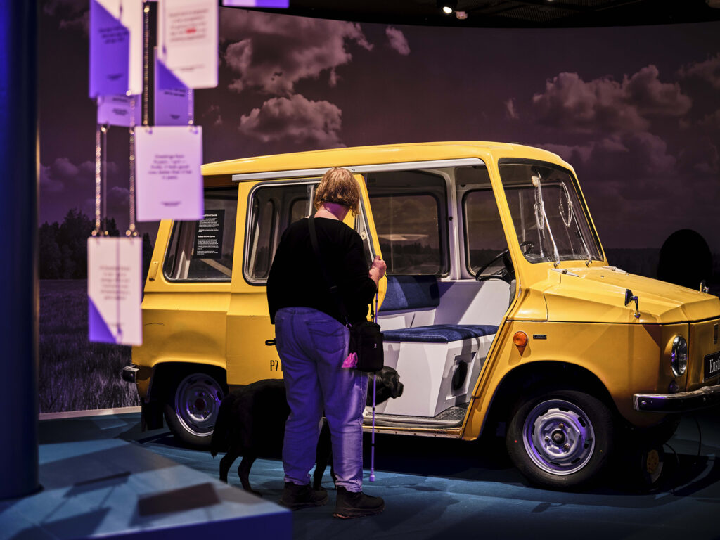 Näyttelytilassa keltainen vanha auto, jonka edessä ihminen valkoisen kepin ja opaskoiran kanssa.