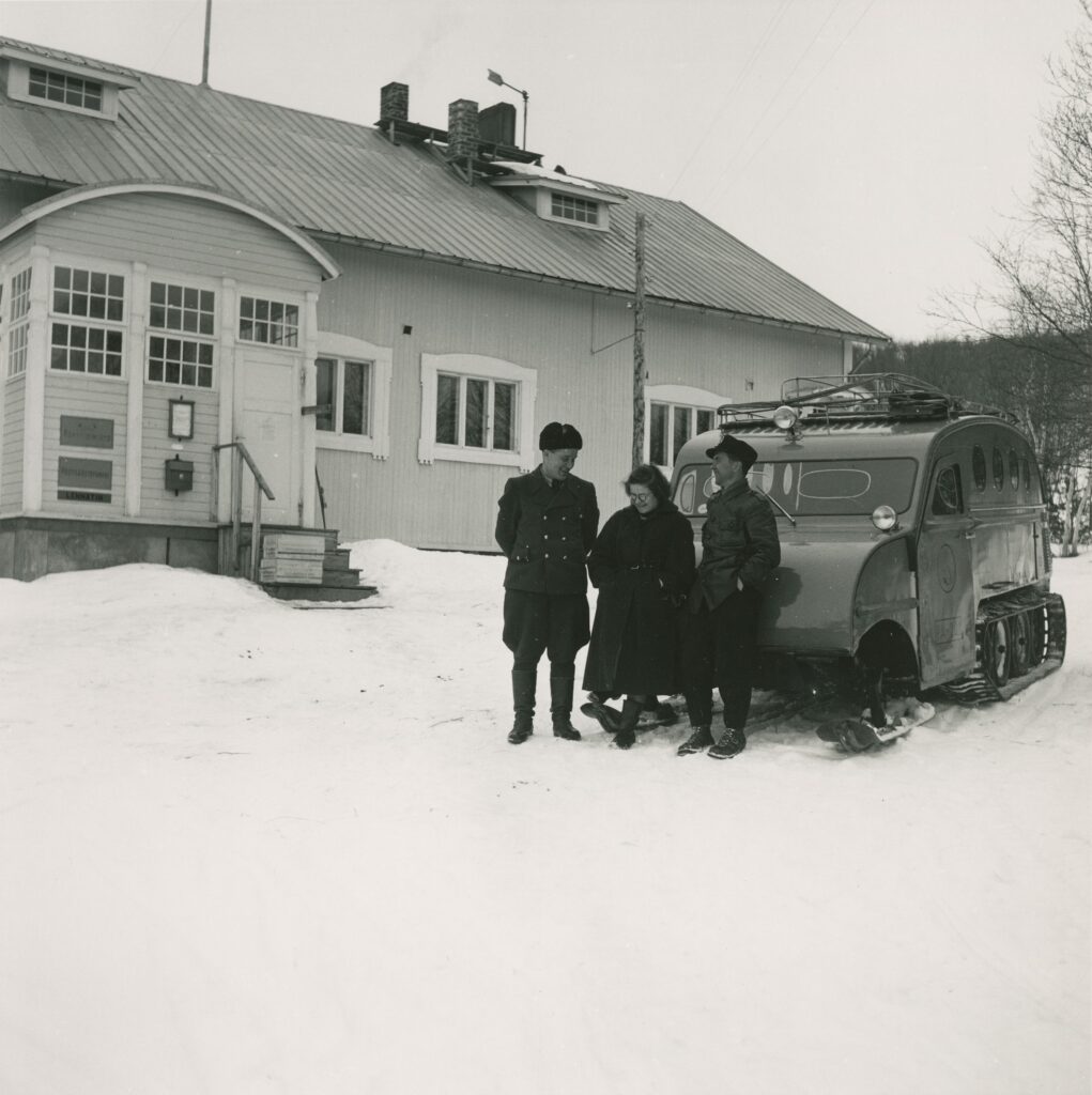 Mustavalkoinen kuva, jossa puinen postitalorakennus taustalla ja lumikiitäjä sekä kolme henkilöä kiitäjän edustalla. Maassa on lunta.