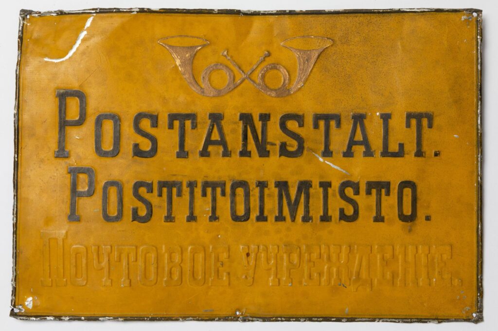 Värivalokuva keltaisesta postitoimipaikkakilvestä, jossa on suomen- ja ruotsinkielinen musta teksti. niiden alla on ollut myös venäjänkielinen teksti, joka on peitetty keltaisella maalilla.