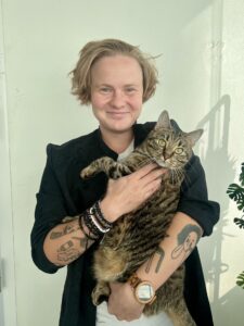 Mies mustassa takissa hymyilee kameralle ja pitää kissaa sylissään, käsissä tatuointeja.
